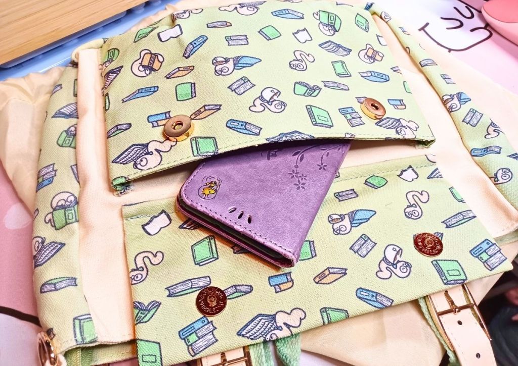 Open pocket in front of a bag. The pocket has a design of cute worms reading books on a green background, and protruding from the pocket is a thick cellphone case.
