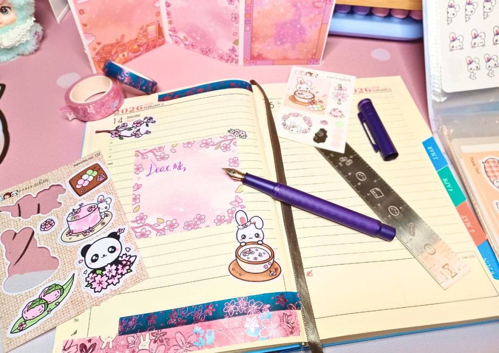 A journal page with cherry blossom washi tapes and sticky notes and adorable animals, with stationery items strewn around like a purple fountain pen and a silver ruler.