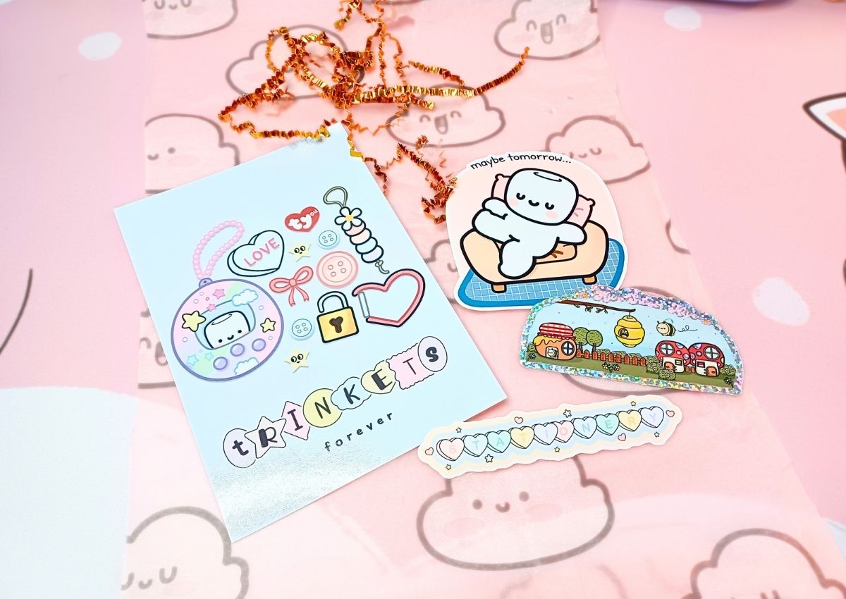 A blue postcard with cute items on it and the word "Trinkets", and three die-cut stickers. One is a cute character lounging on his couch with the words "Maybe tomorrow" above him, one is the word "Stationery" spelled in pastel-colored hearts, and one is a cute mushroom and beehive town on foil paper and the words "Hive Sweet Hive" above it.