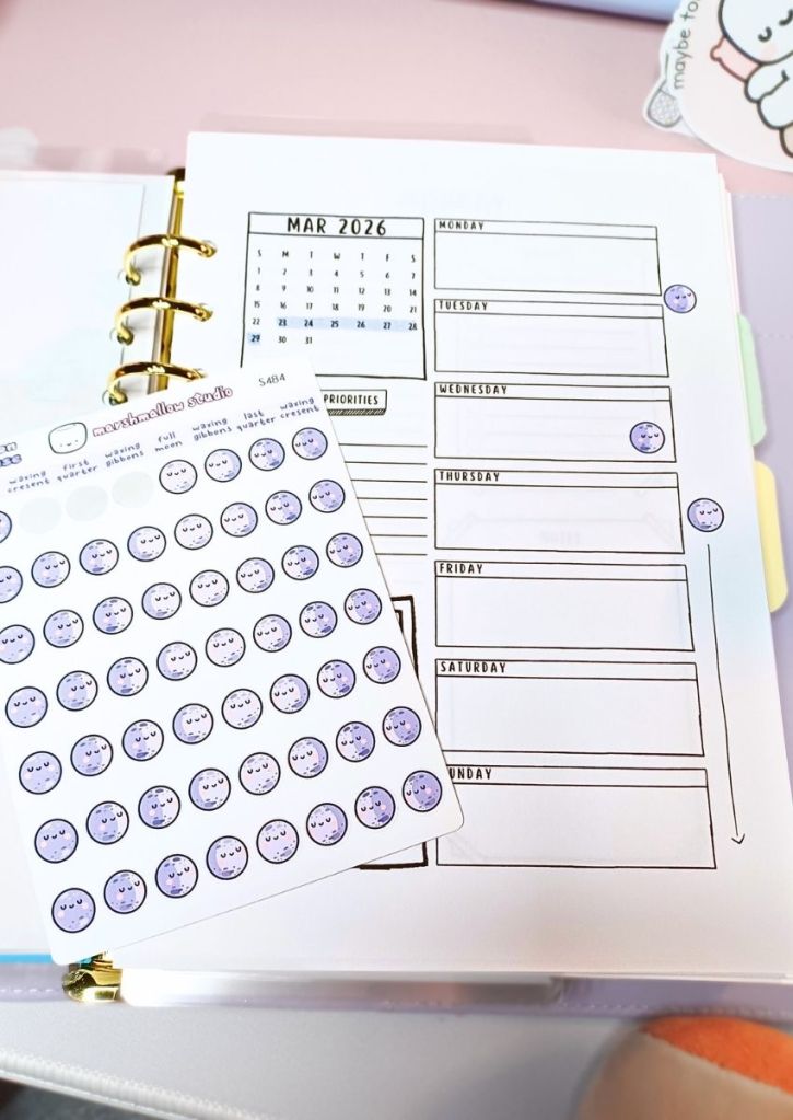 A cute purple moons sticker sheet with some of its stickers removed and stuck to a planner page to mark moon phases.