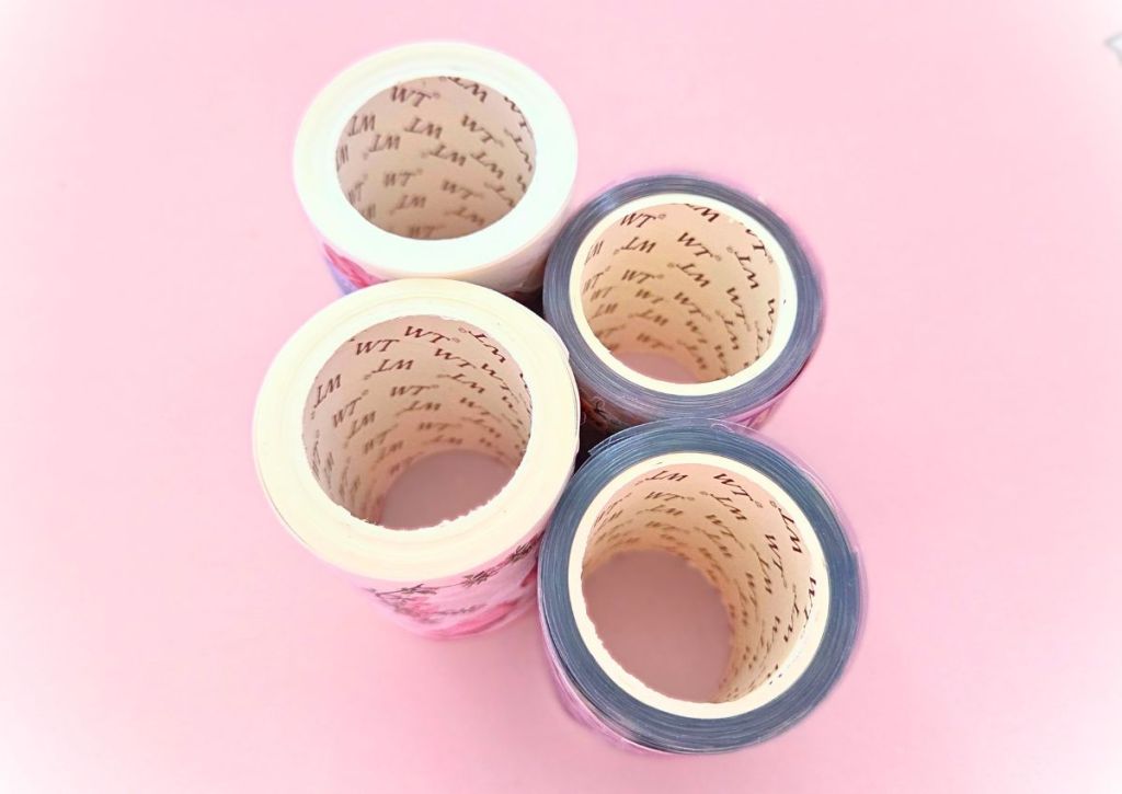 washi-tape-shop-washi-vs-pet Four rolls of decorative washi tape are arranged in a square pattern on a pink background, viewed from above.