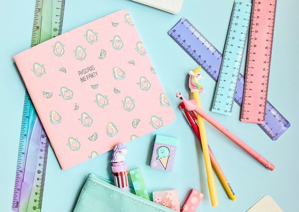 Pink avocado-themed notebook, colorful rulers, unicorn pens, pastel erasers, and a light blue pencil case on a blue surface. The stationery items are playful and arranged neatly, evoking a fun, creative workspace.