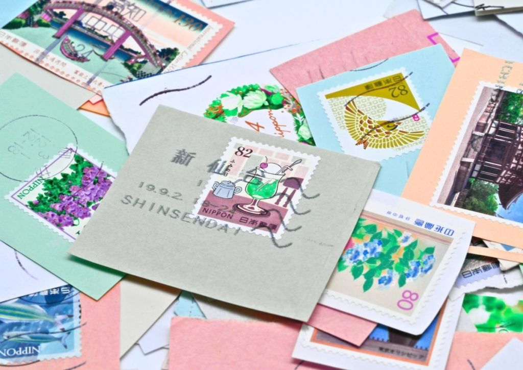	
A variety of colorful Japanese postage stamps are scattered on a surface, featuring flowers, bridges, and traditional art, with one prominently stamped envelope in the center.