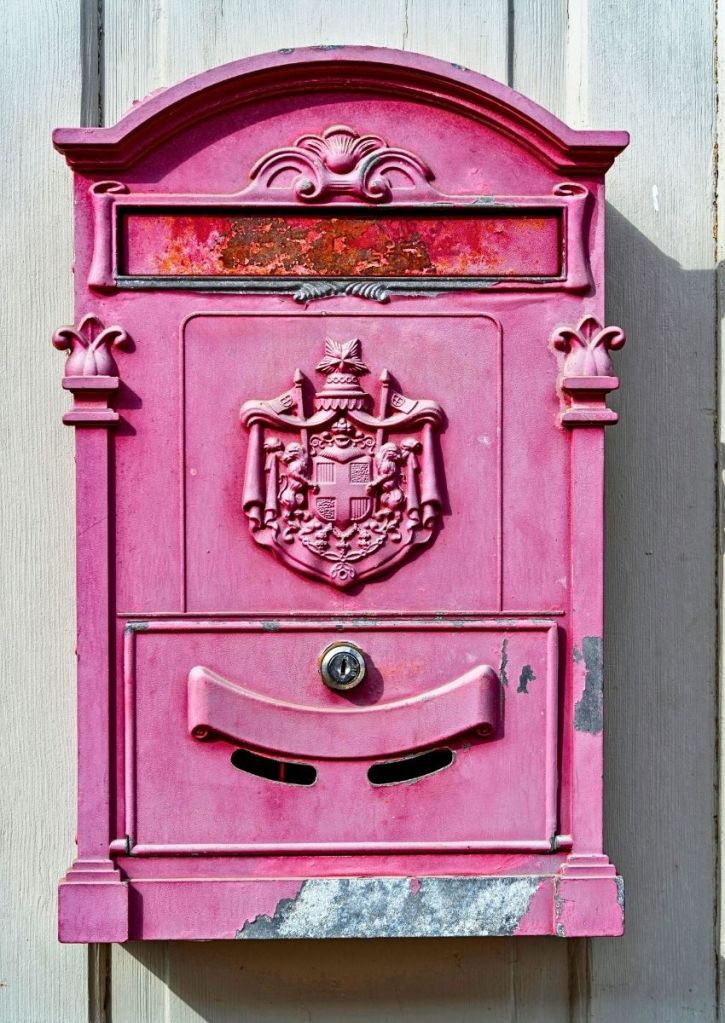 A weathered pink metal mailbox with decorative details and a crest on the front, mounted on a light-colored wooden wall.