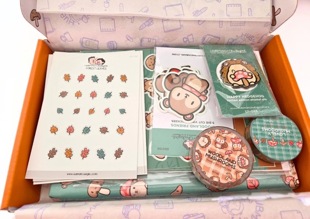 A box filled with cute stationery items, including leaf stickers, cartoon animal stickers, and an enamel pin badge, all with adorable animal and mushroom designs.