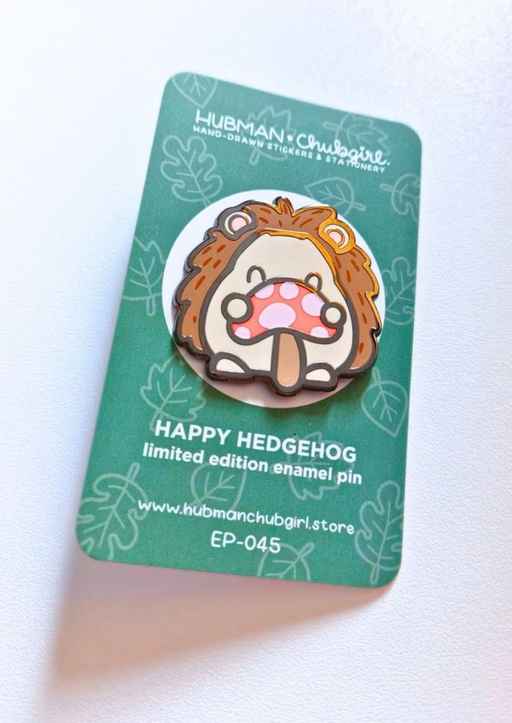 A cute enamel pin of a happy hedgehog holding a mushroom, attached to a green backing card labeled Happy Hedgehog limited edition enamel pin from Hubman & Chubgirl.