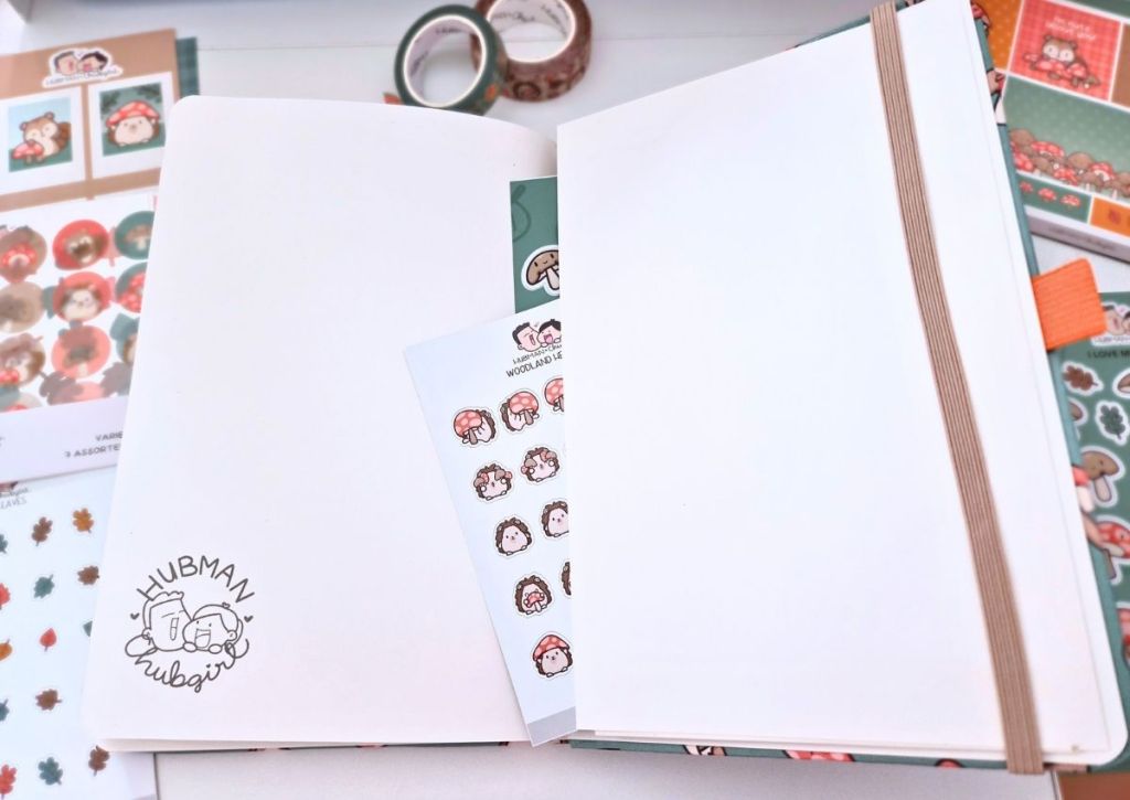 Open notebook with blank pages, elastic band, and cute animal stickers. Surrounding the planner are more sticker sheets, washi tape, and stationery in green, orange, and red tones. Hubman & Chubgirl logo is visible.