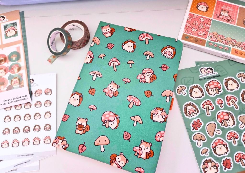 A desk displays cute forest-themed stationery with mushroom and animal designs, including a green notebook, sticker sheets, and washi tape, all featuring matching whimsical illustrations.