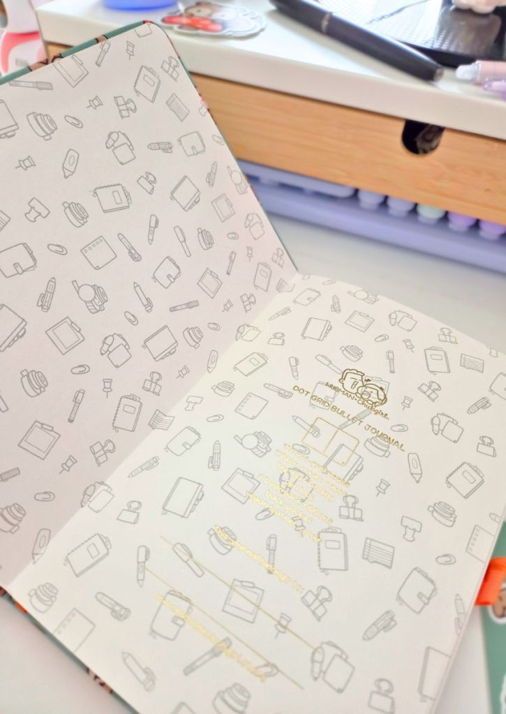 An open journal with patterned pages featuring stationery icons, resting on a desk with stationery items and a drawer visible in the background.