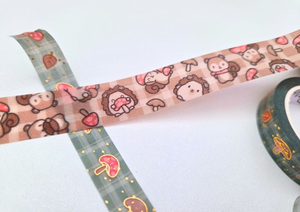 Two rolls of decorative tape: one green with red mushrooms and gold stars, the other beige with illustrations of cute hedgehogs, squirrels, mushrooms, and acorns. The tapes are unrolled and overlap on a light surface.