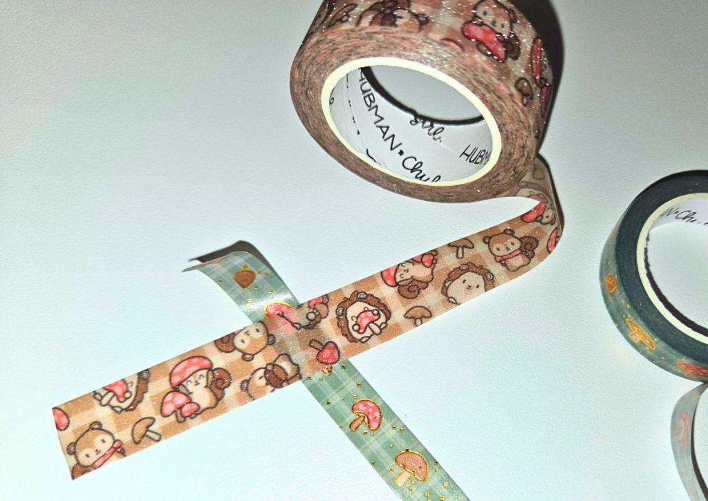 Two rolls of decorative washi tape, one unspooled with a pattern of cartoon hedgehogs, and another with mushrooms. The tapes are arranged on a white surface.