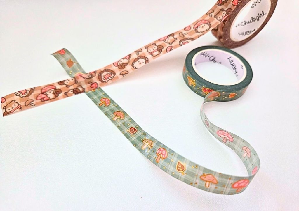 Two rolls of decorative washi tape are unspooled on a white surface. One tape features cartoon animal faces, while the other displays a pattern of mushrooms and leaves.