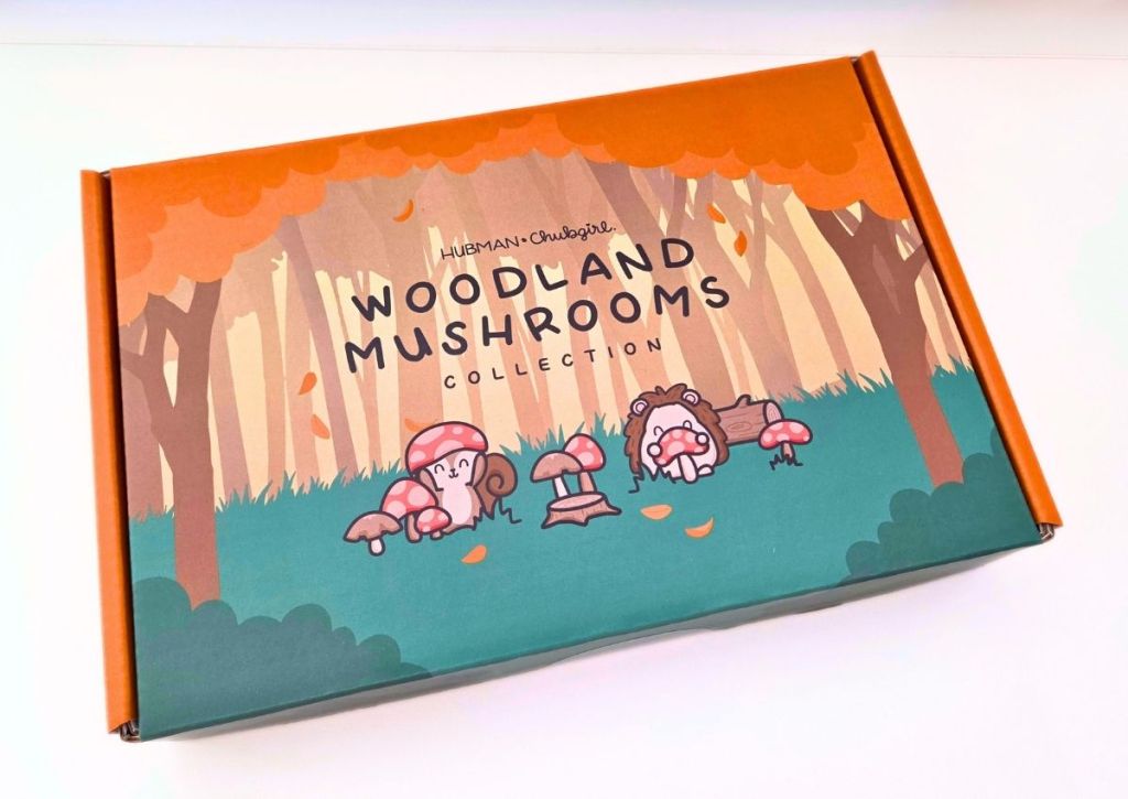 A colorful box with cartoon mushrooms and animals in a forest, labeled WOODLAND MUSHROOMS COLLECTION by Hubman & Chubgirl, on an orange and green background with falling leaves.