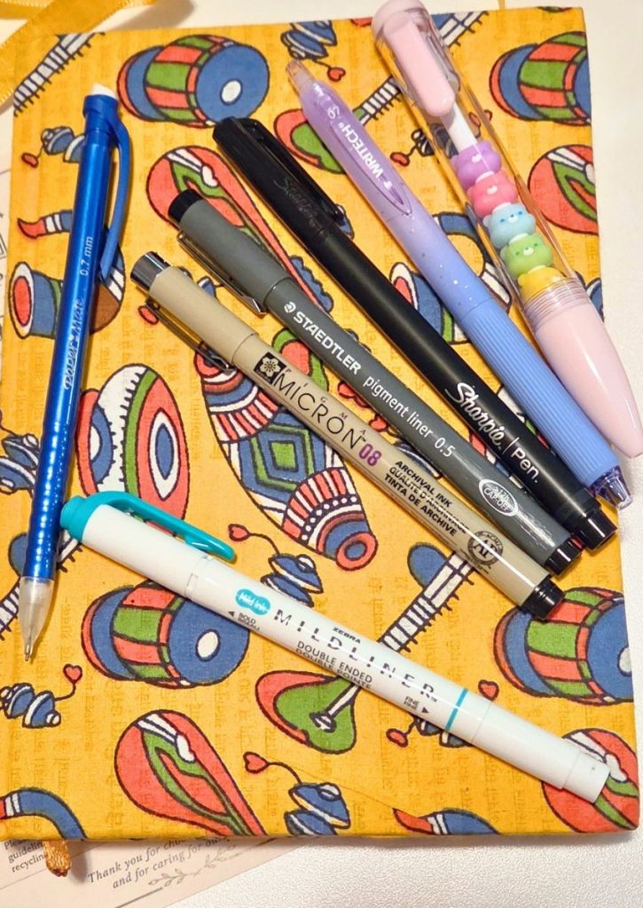 A collection of pens and markers, including a blue mechanical pencil, a black liner, a fine-liner, a black felt-tip pen, and a white double-ended highlighter, resting on a patterned yellow fabric.