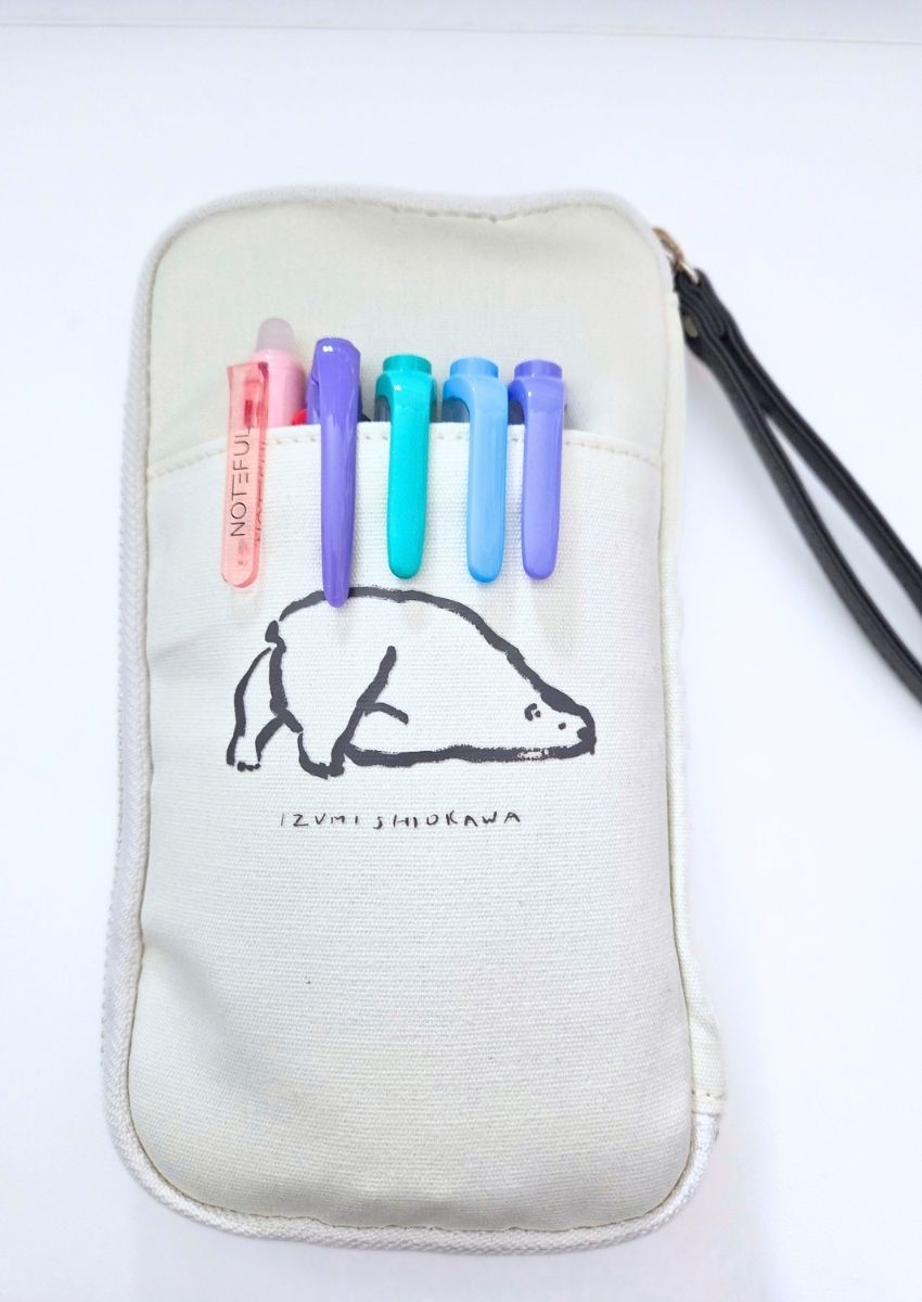 A white canvas pencil case with a black line drawing of a bear and the text Izumi Shiokawa, containing five colorful pens in its front pocket.