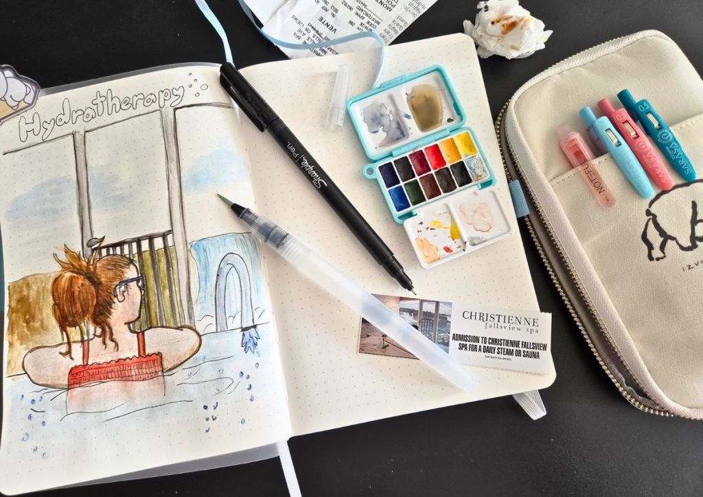 An open bullet journal with a watercolor drawing of a woman in a spa, alongside a black felt-tip pen, water brush, small watercolor set, a spa gift certificate, and a pencil case with gel pens.