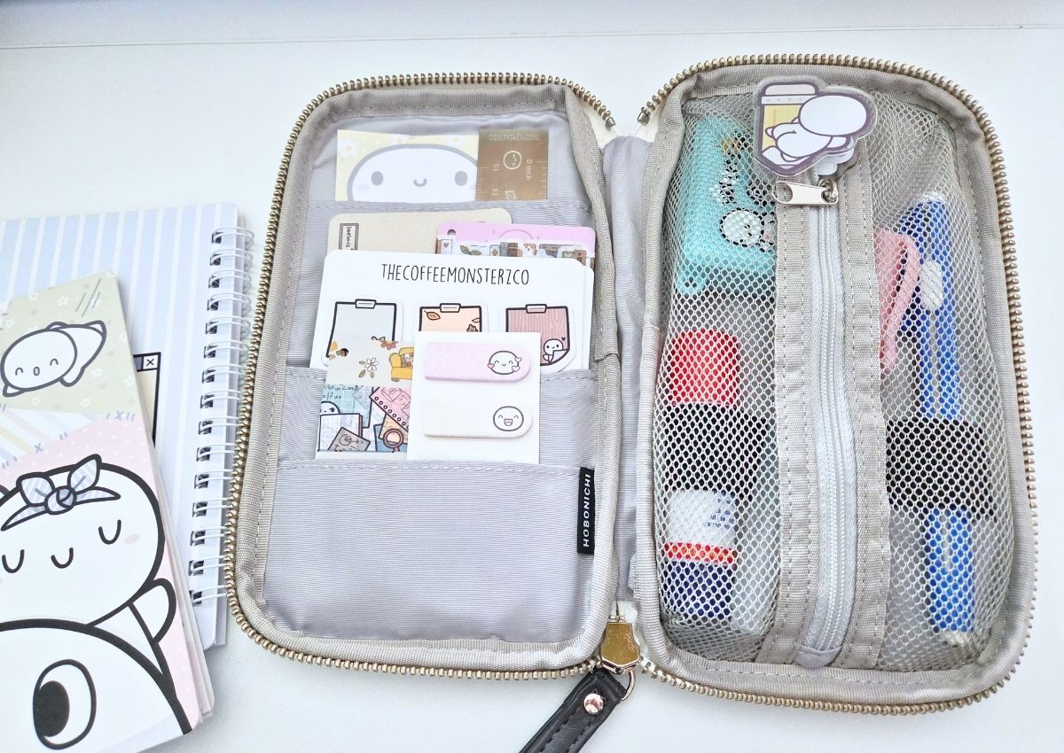 An open zippered organizer case containing stationery, stickers, and cards, sitting next to a spiral-bound notebook.