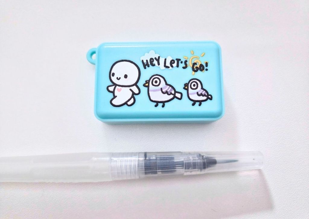 A light blue rectangular case featuring an illustration of a character and two birds with the text Hey Lets Go!, next to a clear water brush pen.