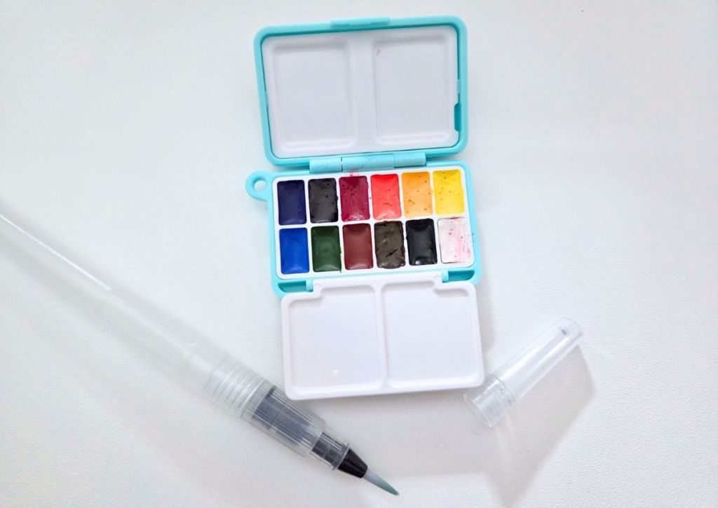 A small, light blue travel watercolor set with twelve paint pans and a water brush pen on a white surface.