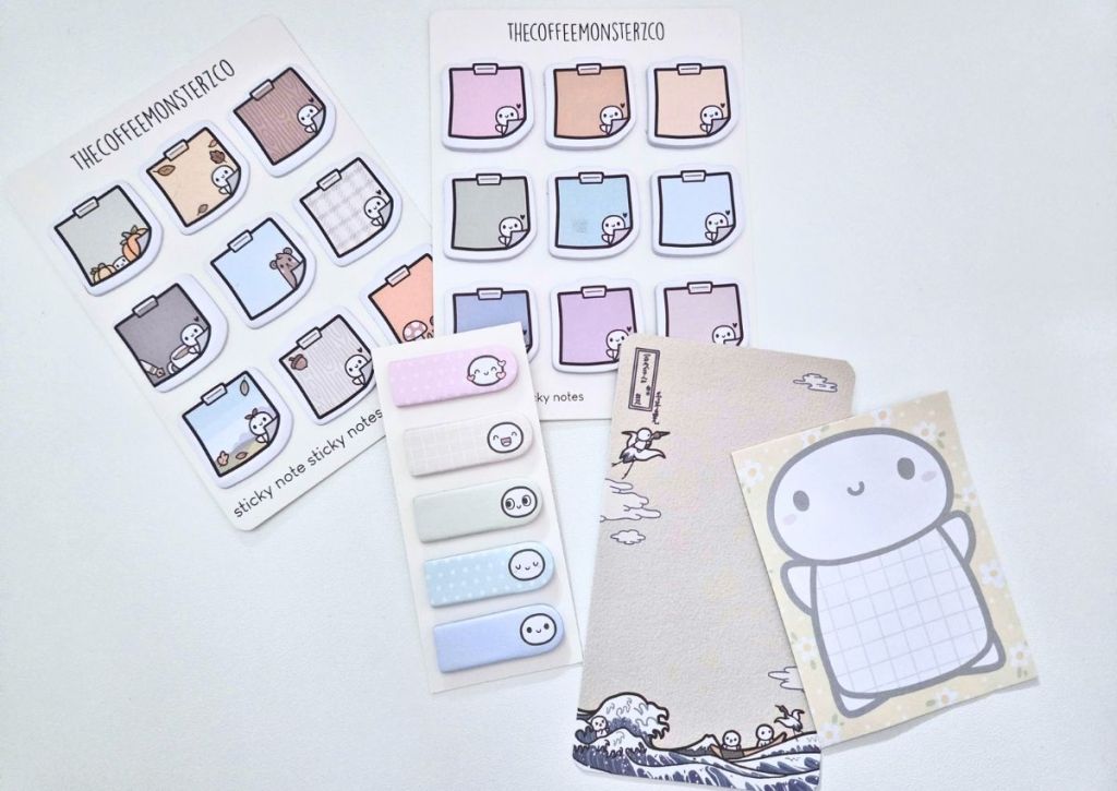 Two sheets of sticky note stickers, a strip of small tab stickers, a Japanese-style illustrated notepad, and a single square notepad featuring a cartoon character, all branded by TheCoffeeMonsterCo.