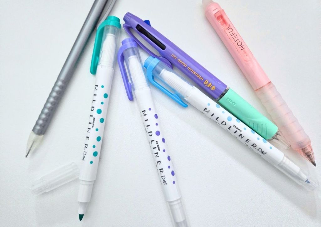 A collection of writing instruments including three Zebra Mildliner Dot pens, a purple Uni pen, and a pink Noteful pen scattered on a white surface.