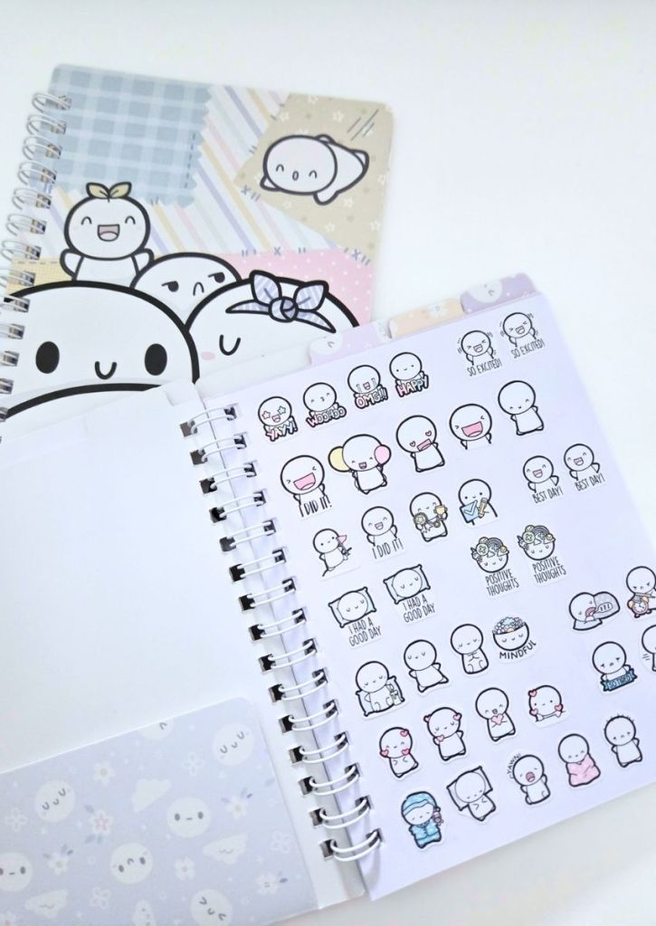 A spiral-bound planner next to a sheet of stickers featuring cute, expressive cartoon characters with text like Yay!, Best day!, and Mindful.