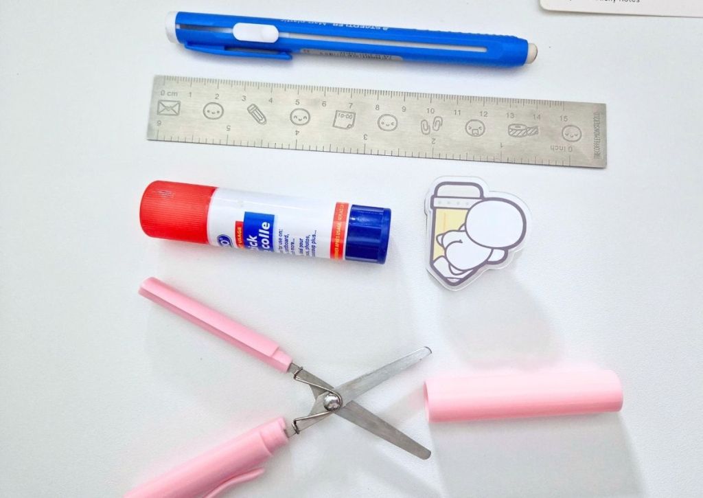 A blue eraser pen, a metal ruler, a glue stick, a cartoon acrylic clip, and a pair of small pink craft scissors on a white surface.