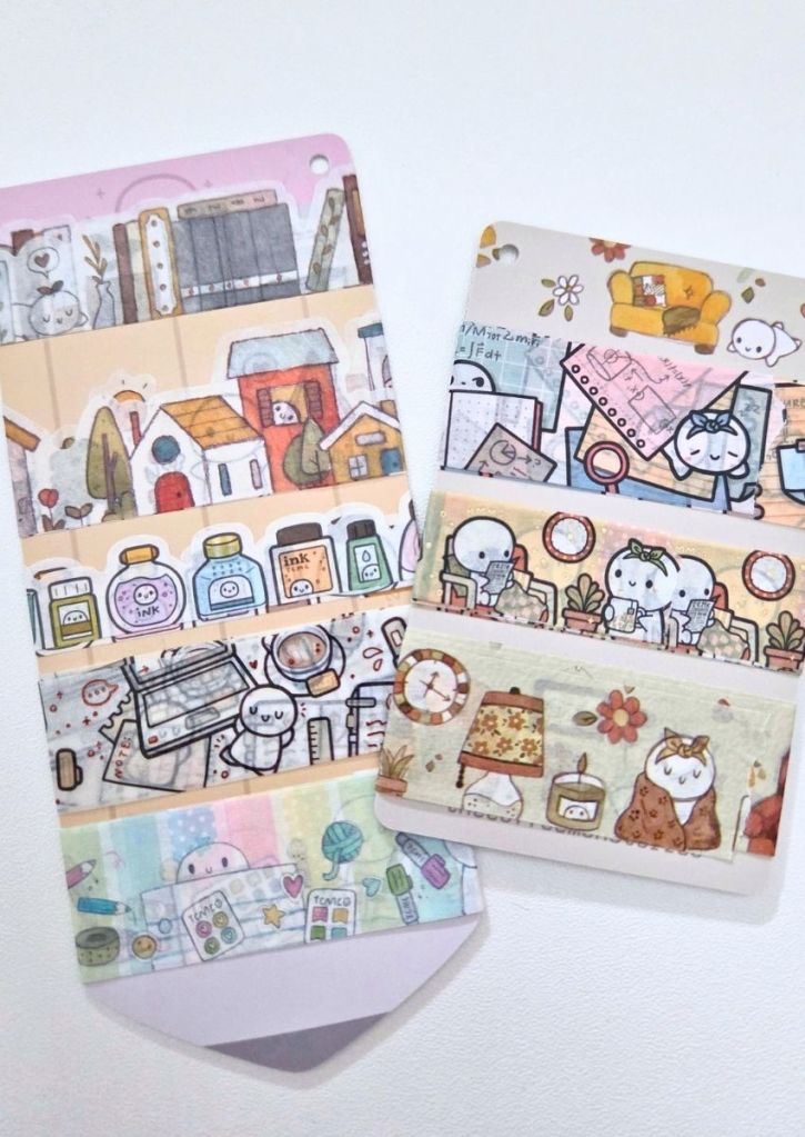 Two sample cards displaying strips of decorative washi tape featuring illustrated scenes of cozy home life, bookshelves, ink bottles, and cute cartoon characters.