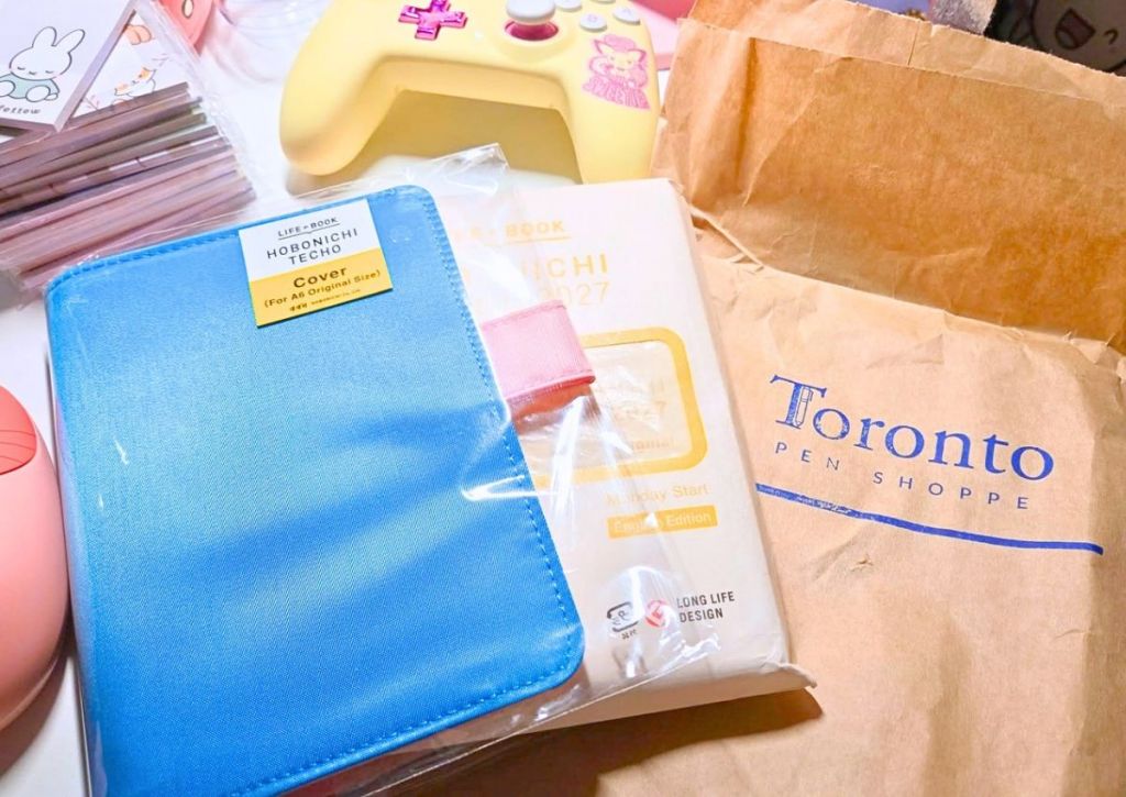 A blue Hobonichi Techo A6 cover, a 2027 Hobonichi planner, and a Toronto Pen Shoppe paper bag.