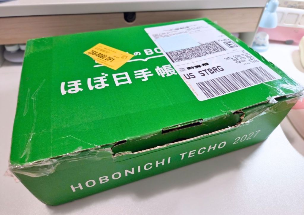 A green Hobonichi Techo 2027 shipping box with a yellow sticker and a shipping label attached.
