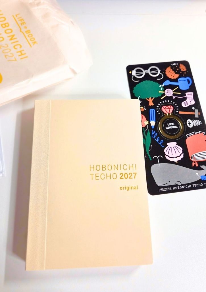 A beige Hobonichi Techo 2027 planner next to a black illustrated bookmark.