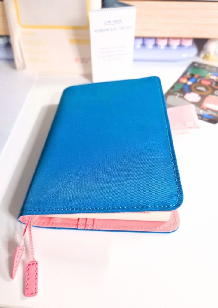 A blue planner cover with pink interior lining and two pink ribbon bookmarks resting on a white desk.