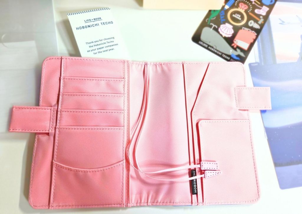 An open, light pink fabric planner cover with interior storage slots, two ribbon bookmarks, and two pen loops.