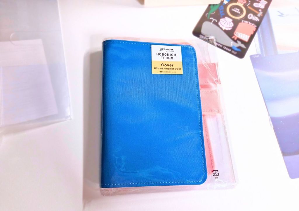 A blue Hobonichi Techo A6 planner cover in plastic packaging, with a decorative bookmark nearby.