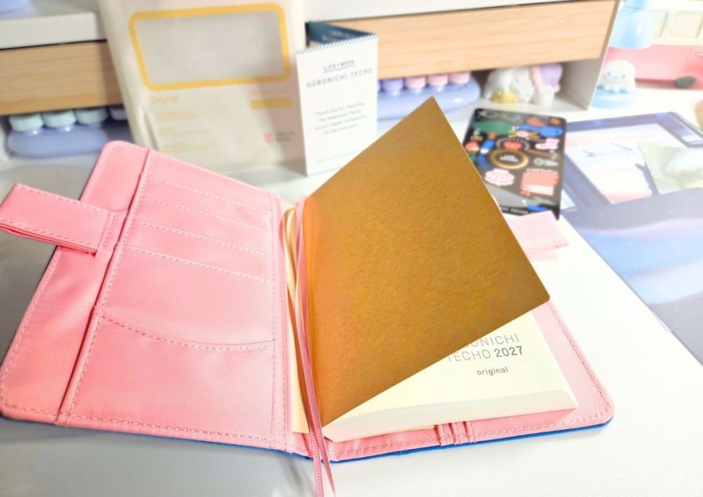 An open pink planner cover with a brown placeholder sheet covering the Hobonichi Techo 2027 planner inside.