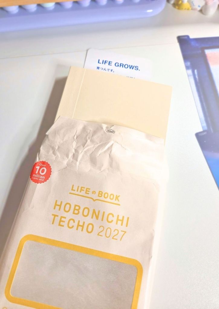 A Hobonichi Techo 2027 planner in its paper packaging, with a sticker reading LIFE GROWS. visible at the top.