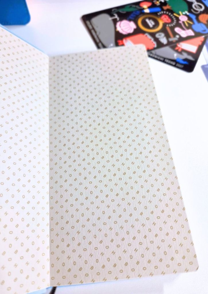 An open notebook with pages patterned in a repeating HOBO text design, with a sticker sheet visible in the background.